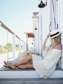 Woman relaxing on wooden steps in a cream knit dress, accessorized with a straw hat and woven bag.