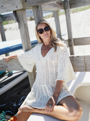 Woman seated near wooden dock beams in a cream knit dress, wearing oversized sunglasses and enjoying a sunny day.