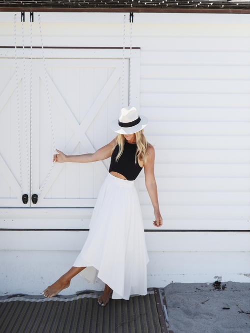 Woman wearing a white asymmetrical skirt and black tank top, playfully holding a hat, with a white structure in the background.