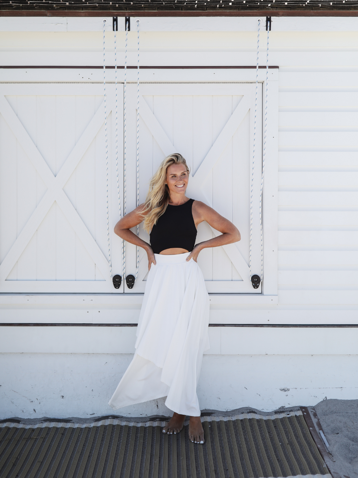 Full-length image of a woman wearing a black top and white skirt, smiling confidently in front of a white barn-style door.