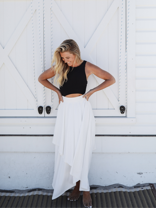 Close-up of a woman wearing a black tank top with a cutout waist and a white skirt, smiling with her hands on her hips, set against a clean, minimalist background.