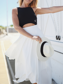 Woman wearing a black tank-style top with a white layered skirt, holding a white sun hat with a black ribbon, set against a bright, sunny backdrop