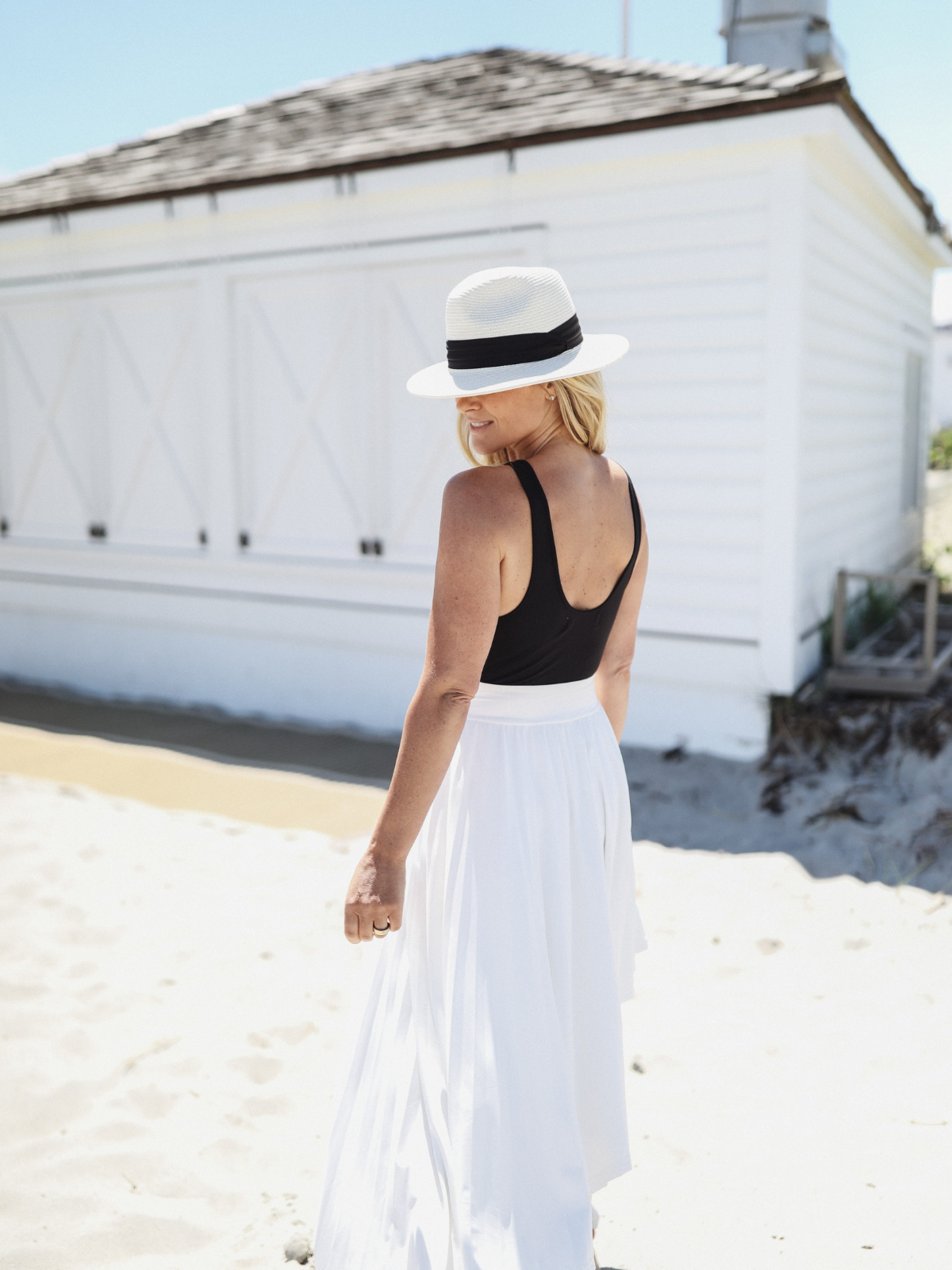 Woman walking along the beach wearing a black sleeveless top with a scoop back and a white skirt, accessorized with a white hat.