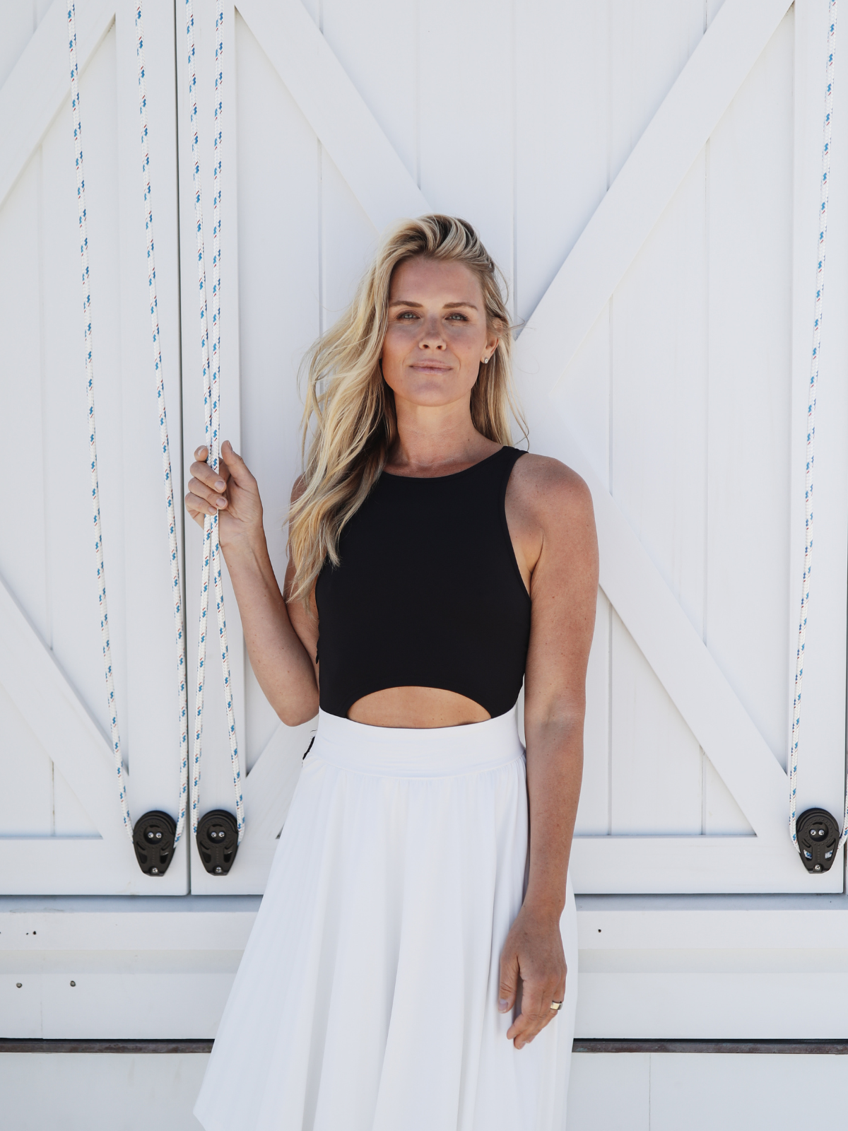 Woman posing in a sleeveless black top with a cutout at the waist, paired with a flowing white asymmetrical skirt, standing against a white wooden door with nautical ropes.