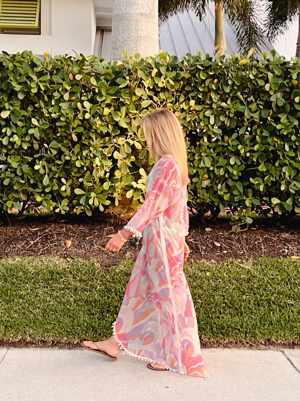 Woman walking along a sidewalk in a flowing colorful caftan with pom-pom detailing, surrounded by greenery.