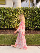 Woman walking along a sidewalk in a flowing colorful caftan with pom-pom detailing, surrounded by greenery.