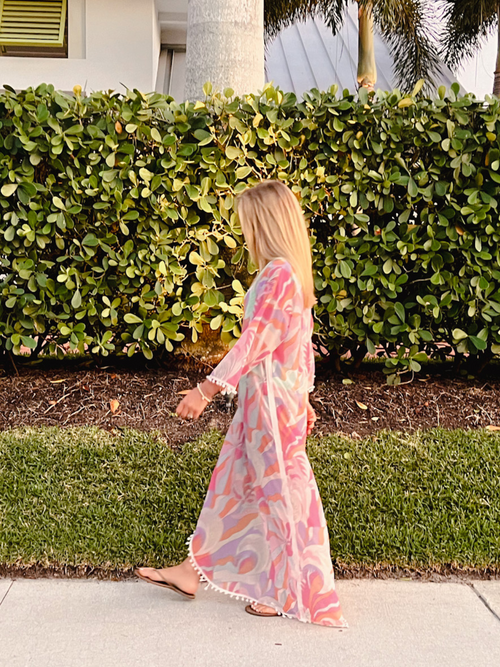 Woman walking along a sidewalk in a flowing colorful caftan with pom-pom detailing, surrounded by greenery.