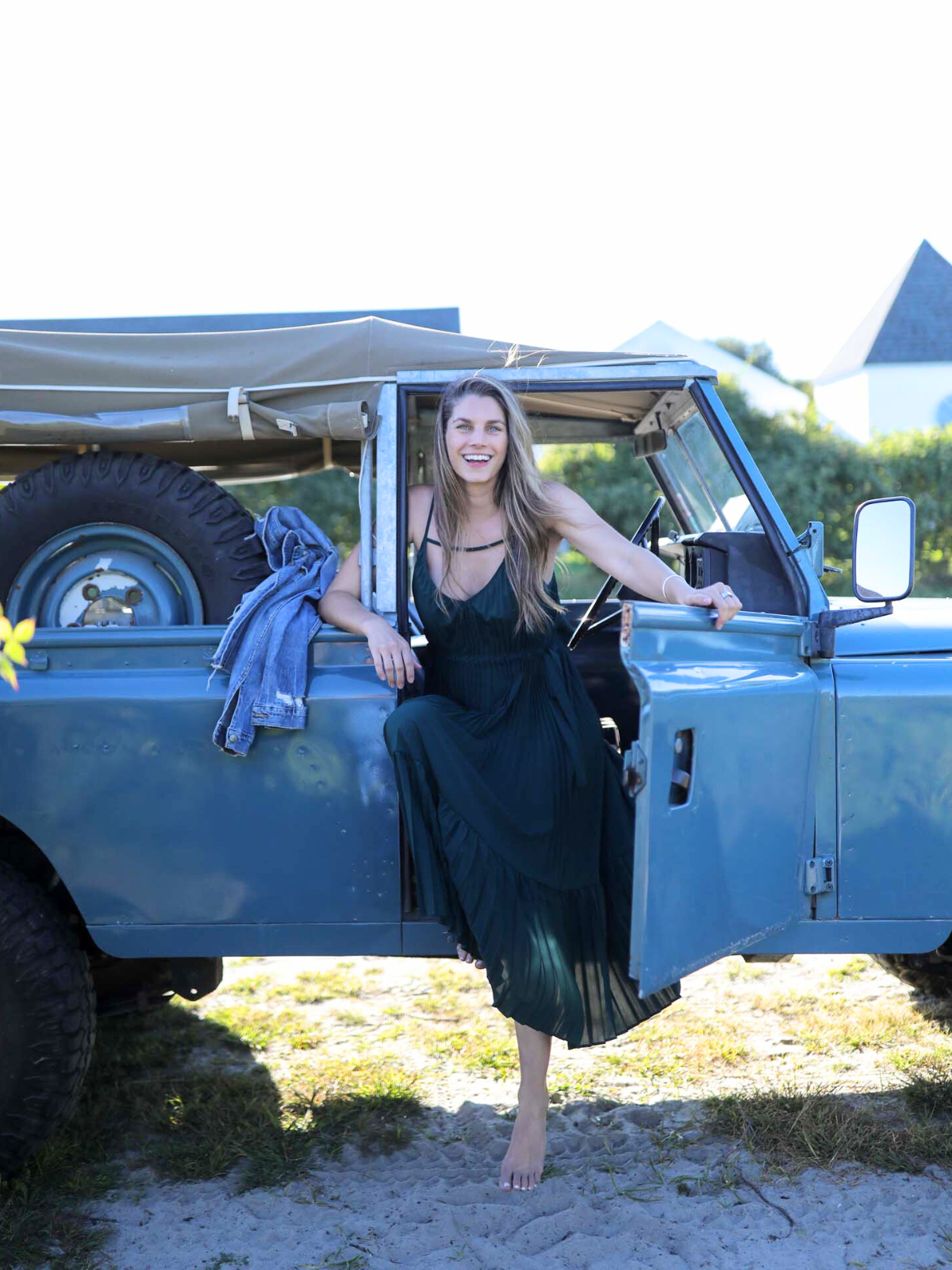 A woman sitting in the open doorway of a vintage blue SUV, wearing a dark green pleated maxi dress, enjoying a relaxed outdoor moment.