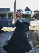 A woman joyfully twirls in a dark green pleated maxi dress, capturing movement and light in a natural outdoor setting.