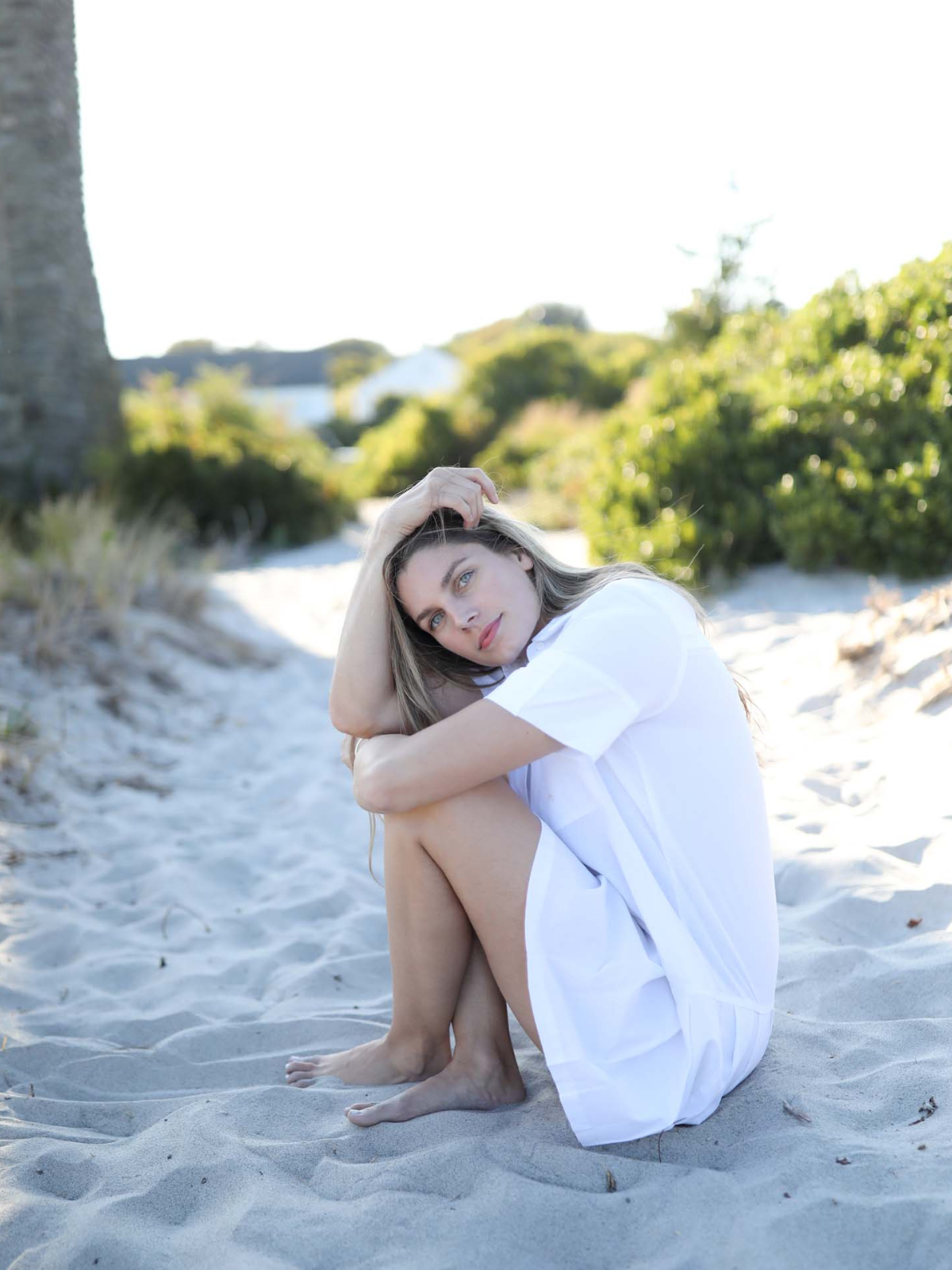 Woman sitting on a sandy beach path with her knees up, wearing a white shirtdress and gazing into the distance.