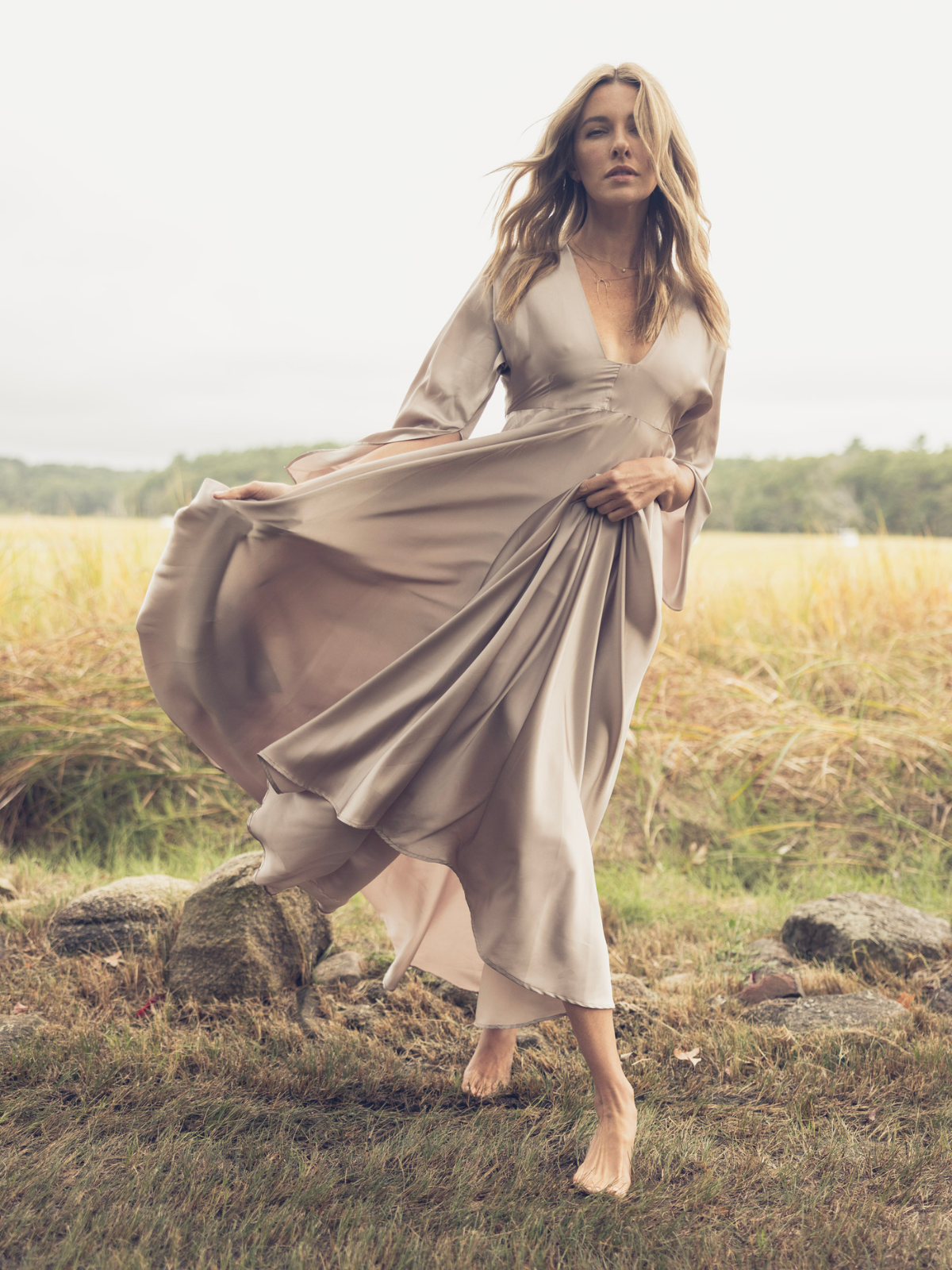 A woman stands in a flowing silky sand-colored maxi dress with long flutter sleeves and a deep V-neck, captured in motion against a grassy field.