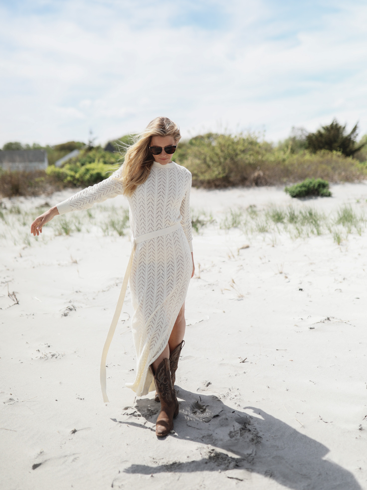 Woman walking on the beach in a cream knit maxi dress with a slit and chevron pattern, paired with brown cowboy boots, under a bright blue sky.