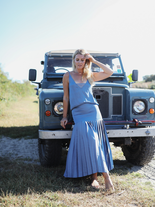 A woman standing in front of a vintage Land Rover, wearing a blue cami and a matching pleated skirt, exuding casual elegance.