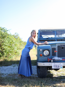 A serene shot of a woman leaning against a vintage Land Rover, wearing a fitted blue cami and pleated skirt, set against a natural backdrop.